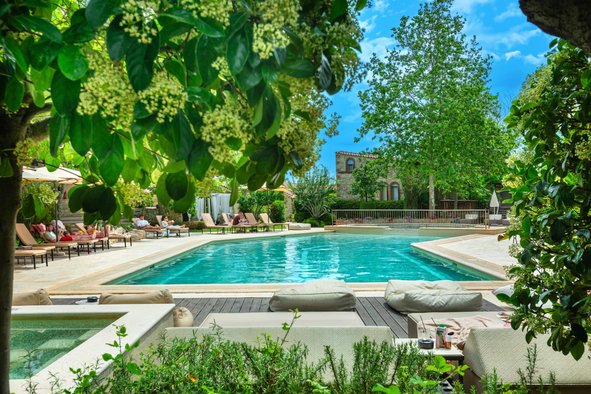The main pool and jacuzzi surrounded by sun loungers, tall trees and stone houses.